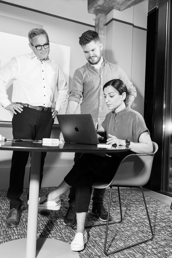 three people standing or sitting around table with laptop