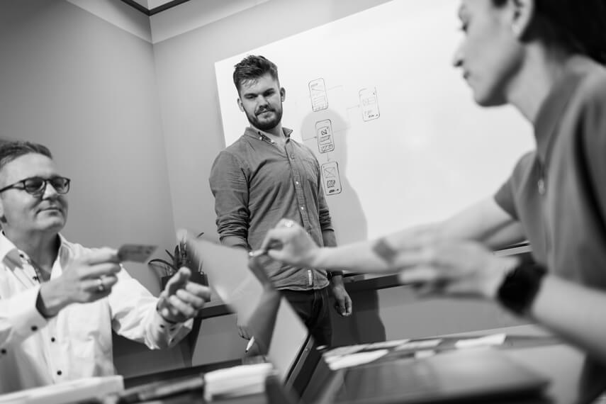 2 persons giving something to each other in a meeting, a third person is watching them and standing next to a whiteboard.