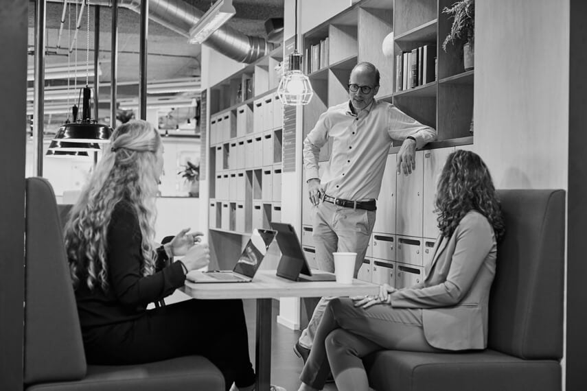 2 women sitting on couch with a table inbetween, talking to a man standing next to them.