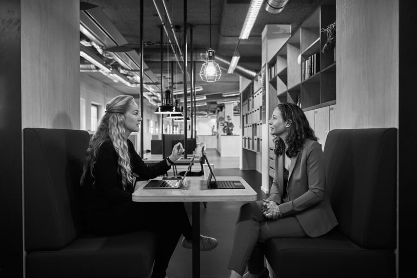 two woman sitting at table with laptops and talking