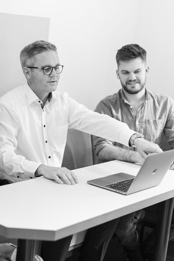 two men sitting at a table looking at a laptop.